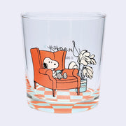 Clear glass with Snoopy design sitting on an orange chair next to a plant atop of a checkered floor.