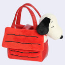 A red plush handbag styled like Snoopy's house, featuring black stitched lines and a plush Snoopy head peeking out from the side, set against a light gray background.