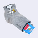 Gray ankle socks with a graphic of Snoopy and Woodstock walking with one another on the ankle part of the sock. At the toe, there is a white and black stripe.