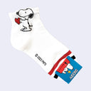 A single white ankle sock featuring a cartoon Snoopy design, with Snoopy holding a red heart near the cuff. The sock has a red stripe, a blue cuff label with Snoopy graphics, and a visible copyright mark reading 2023 PNTS.
