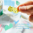 A close-up of a hand holding two small yellow cat-face stickers above a transparent sheet of assorted decorative stickers. The scene is brightly lit with soft-focus background elements and Japanese text overlaid in the lower right.