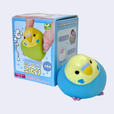 Small figurine of a blue and yellow parakeet bird, round and chubby. It sits next to its product packaging.