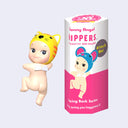 Vinyl figure of a Kewpie baby, nude aside from a yellow cat shaped hood hat. It looks back over its shoulder with one leg hoisted up, as though climbing something.