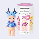 A Sonny Angel x shu uemura Makeup Angel Series collectible figure stands next to its product packaging box. The figure is wearing a shiny blue deer-antler hooded sweatshirt with a pale peach body.