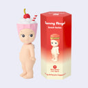 Sonny Angel mini figure - a nude Kewpie baby standing with a cute smile and a strawberry milkshake atop its head, like a hood hat. It stands next to its product packaging.