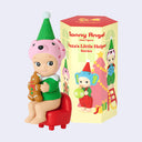 Vinyl figure of a Sonny Angel, wearing a green Christmas elf outfit and sitting on a red chair. It holds a gingerbread man and has a pink Dalmatian hat in front of its product packaging. 