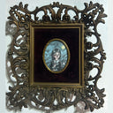 Illustration in a small oval within a black velvet mat and ornate bronze frame. Illustration is of a small girl with braids and a fancy collared dress. She looks off to the side while smiling.