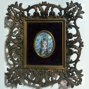 Illustration in a small oval within a black velvet mat and ornate bronze frame. Illustration is of a small girl with braids and a fancy collared dress. She looks off to the side while smiling.