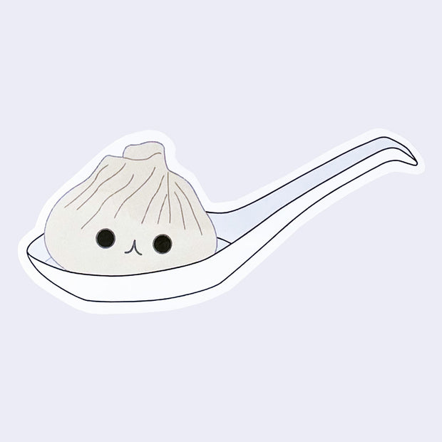 Die cut sticker of an illustrated soup dumpling in a large ramen spoon. Dumpling has a smiling face.