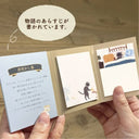 A person holds an open small booklet with Japanese text and illustrated panels, including a black cat illustration, against a wooden floor background. A cream speech-bubble overlay contains Japanese text.