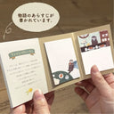 Small book or notebook held open by a hand, showing illustrations and text on a wooden surface.