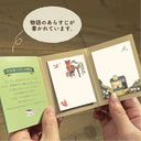 Small book with illustrated pages held by hands, featuring a house and animal illustrations.