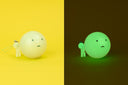 Promo graphic for Smiski: Strap Accessories capsule with a photo shown in light and in dark, of a small Smiski next to a larger capsule of Smiski's head - both glowing in the dark.