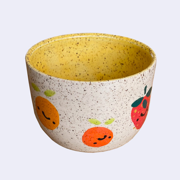 Ceramic bowl with spotted finishing and an earthy cream exterior and mustard yellow interior. On the outside are painted on cartoon style citrus and a strawberry, with simple expressions.