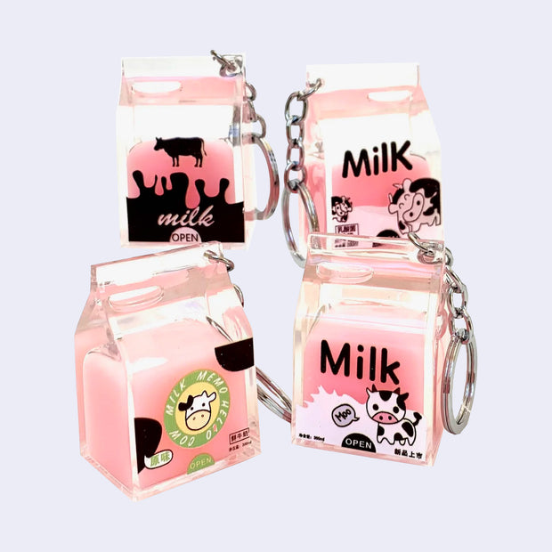 4 plastic keychains in the shape of a milk carton, transparent and containing a pink liquid. Each carton has a different front design, featuring the words "milk" and a graphic of a cow. 