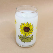 Glass cup with a flat base and slightly inward lip. Features a graphic of a cartoon style sunflower, sitting with a smiling face and a chubby green body made out of stems and leaves. Around the rest of the glass are yellow sunflowers.