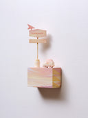 Small rectangular wood panel painted like a pinkish orange sunset with a mountain range barely seen at the very bottom. Atop of it are carved wooden sculptures: a retro looking pink and orange car and a pole with large signage, similarly colored with the word "sunseeker" on it.