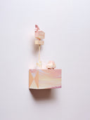 Small rectangular wood panel painted like a pinkish orange sunset with a mountain range barely seen at the very bottom. Atop of it are carved wooden sculptures: a retro looking pink and orange car and a pole with large signage, similarly colored with the word "sunseeker" on it.