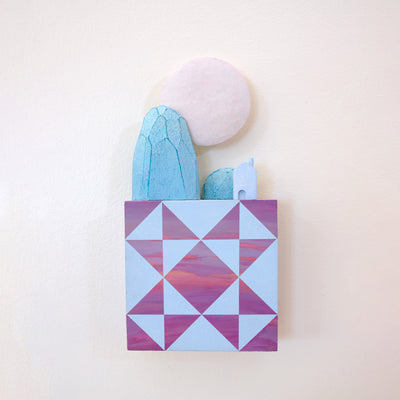 Small square wood panel painted like a pinkish purple sunset with light blue patterning overlayed on top, akin to a quilt tile design. Atop of it are carved wooden sculptures: blue mountainous rocks, a small blue house and a large white moon.