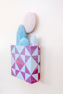 Small square wood panel painted like a pinkish purple sunset with light blue patterning overlayed on top, akin to a quilt tile design. Atop of it are carved wooden sculptures: blue mountainous rocks, a small blue house and a large white moon.