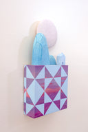 Small square wood panel painted like a pinkish purple sunset with light blue patterning overlayed on top, akin to a quilt tile design. Atop of it are carved wooden sculptures: blue mountainous rocks, a small blue house and a large white moon.