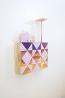 Small square wood panel painted like an orange-purple sunset with light purple patterning overlayed on top, akin to a quilt tile design. Atop of it are carved wooden sculptures: 3 small pastel colored houses and a tall flower.