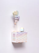 Small rectangular wood panel painted like a multicolor sunset with white triangle patterning overlayed on top, akin to a quilt tile design. Atop of it are carved wooden sculptures: a small, retro looking blue van bus and 2 posts with several signs attached, painted yellow, blue and purple.