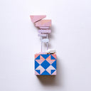 Small square wood panel painted like a multicolor sunset with bright blue patterning overlayed on top, akin to a quilt tile design. Atop of it are carved wooden sculptures: a small, retro looking blue car and 2 posts with several signs attached, painted in pinkish sunset tones.