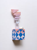 Small square wood panel painted like a multicolor sunset with bright blue patterning overlayed on top, akin to a quilt tile design. Atop of it are carved wooden sculptures: a small, retro looking blue car and 2 posts with several signs attached, painted in pinkish sunset tones.