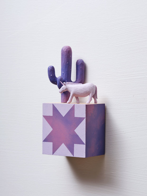 Small square wood panel painted like a purple sunset with light pink patterning overlayed on top, akin to a quilt tile design. Atop of it are carved wooden sculptures: a purple Saguaro cactus and a pinkish purple unicorn.