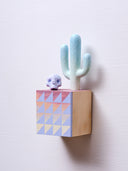 Small square wood panel painted like an orange sunset with light purple triangle patterning overlayed on top, akin to a quilt tile design. Atop of it are carved wooden sculptures: a light mint green Saguaro cactus and a small purple skull.