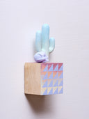 Small square wood panel painted like an orange sunset with light purple triangle patterning overlayed on top, akin to a quilt tile design. Atop of it are carved wooden sculptures: a light mint green Saguaro cactus and a small purple skull.