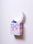 Small square wood panel painted like an purple sunset with light purple triangle patterning overlayed on top, akin to a quilt tile design. Atop of it are carved wooden sculptures: a small blue and purple boat and a large crescent moon.