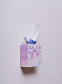 Small square wood panel painted like an purple sunset with light purple triangle patterning overlayed on top, akin to a quilt tile design. Atop of it are carved wooden sculptures: a small blue and purple boat and a large crescent moon.