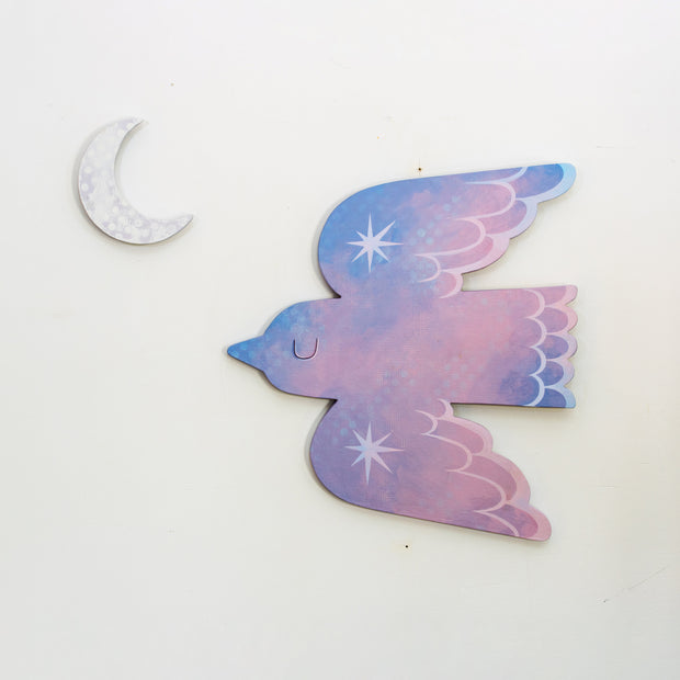 Die cut bird shaped canvas. A pink and blue painted bird flies with an eye closed and sparkles on its wings. A crescent moon hangs nearby.