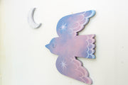 Die cut bird shaped canvas. A pink and blue painted bird flies with an eye closed and sparkles on its wings. A crescent moon hangs nearby.