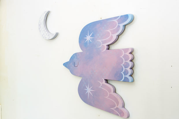 Die cut bird shaped canvas. A pink and blue painted bird flies with an eye closed and sparkles on its wings. A crescent moon hangs nearby.