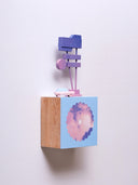 Small square wood panel with a purple and pink sunset shown within a flower outline, the rest of the panel is painted light blue. Atop of it are carved wooden sculptures: a small pink colored retro car and a several large signs attached to 2 poles.