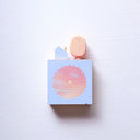 Small square wood panel with an orange and pink sunset shown within a flower outline, the rest of the panel is painted light blue. Atop of it are carved wooden sculptures: a small light blue house and an oval shaped yellow tree.