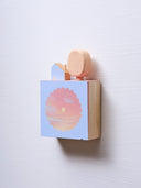 Small square wood panel with an orange and pink sunset shown within a flower outline, the rest of the panel is painted light blue. Atop of it are carved wooden sculptures: a small light blue house and an oval shaped yellow tree.