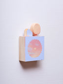 Small square wood panel with an orange and pink sunset shown within a flower outline, the rest of the panel is painted light blue. Atop of it are carved wooden sculptures: a small light blue house and an oval shaped yellow tree.