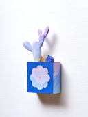 Small square wood panel with an purple and pink sunset shown within a flower outline, the rest of the panel is painted dark blue. Atop of it are carved wooden sculptures: a small dark blue house, a brown rock and a tall purple cactus.