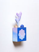 Small square wood panel with an purple and pink sunset shown within a flower outline, the rest of the panel is painted dark blue. Atop of it are carved wooden sculptures: a small dark blue house, a brown rock and a tall purple cactus.