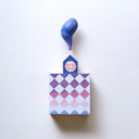 Small square wood panel painted like a pink and purple sunset with light blue diamond patterning overlayed on top, akin to a quilt tile design. Atop of it is a carved wooden house with a large painted flower on the front and a thick plume of smoke coming out the top.