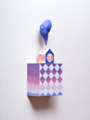 Small square wood panel painted like a pink and purple sunset with light blue diamond patterning overlayed on top, akin to a quilt tile design. Atop of it is a carved wooden house with a large painted flower on the front and a thick plume of smoke coming out the top.