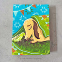 A painting of a tan dog lying under a green blanket next to a green bird, with a bright blue patterned sky and colorful bunting above. The artwork is photographed against a light gray textured surface.
