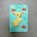 A whimsical illustration of a light yellow dog with pink-tipped ears, surrounded by three small red birds on a turquoise background with white and teal geometric patterns, photographed flat on a gray wood surface.