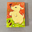A whimsical illustration of a cream-colored poodle on a green patterned ground, with an orange patterned sky and two small dark birds, photographed on a gray textured surface.