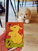 A hand holds a small painting of a yellow dog on a red and green patterned background, with a small fluffy dog blurred in the background on a wooden floor.