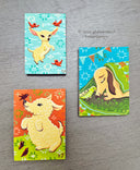 Three colorful paintings featuring cute animals and birds, arranged on a gray wood-textured surface. One card shows a light yellow puppy on an orange background, one shows a light yellow chihuahua on a light blue background, and one shows a light yellow puppy under a green blanket on a blue background.
